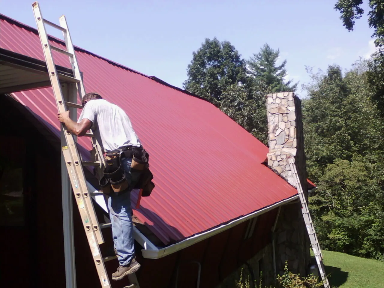 Roofer on a ladder beside a red metal roof and stone chimney — Soffit & Fascia Repair by Roofing Brothers in Easthampton